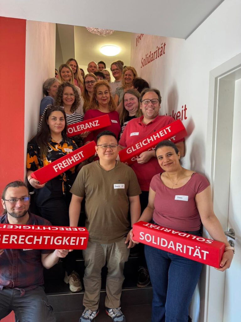 Ein Gruppenfoto mit einigen Mitarbeitern. Fünf Mitarbeiter halten jeweils einen Holzbalken in der Hand mit jeweils einer Aufschrift: Gerechtigkeit, Solidarität, Gleichheit, Freiheit und Toleranz,